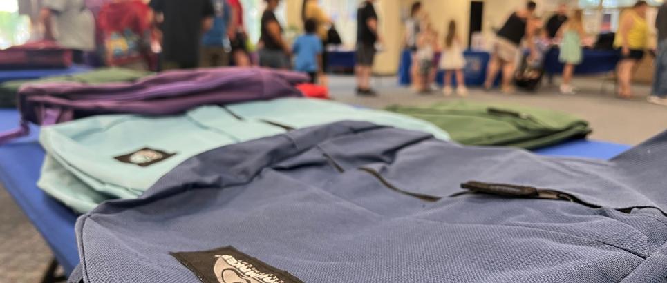 Backpacks lay on a table while people line up at a school supply distribution event