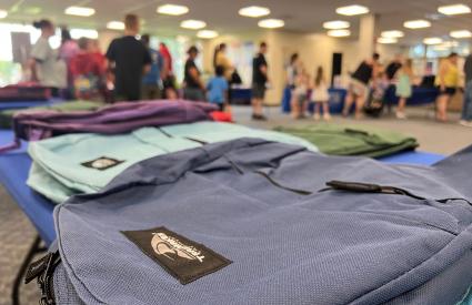 Backpacks lay on a table while people line up at a school supply distribution event