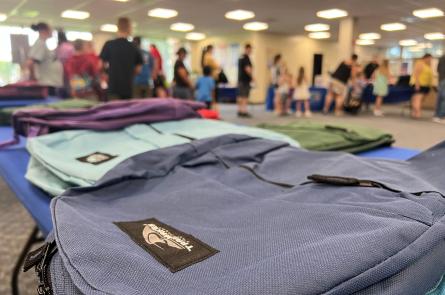 Backpacks lay on a table while people line up at a school supply distribution event