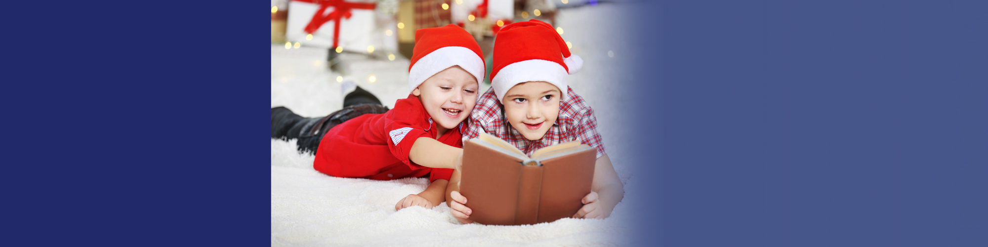 Two kids in santa hats read a book while smiling