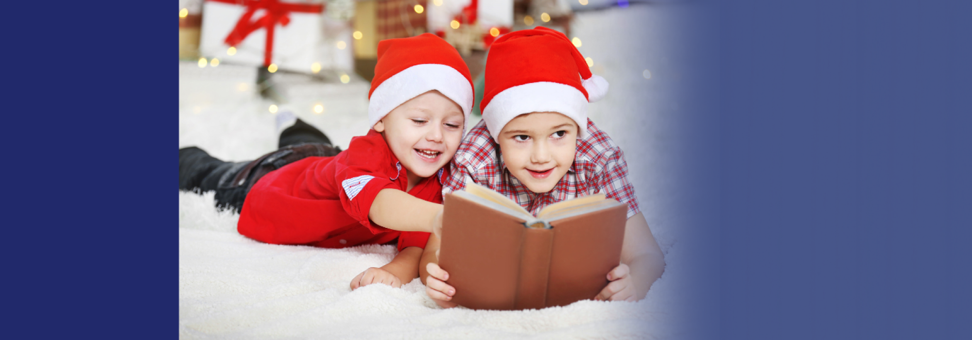 Two kids in santa hats read a book while smiling